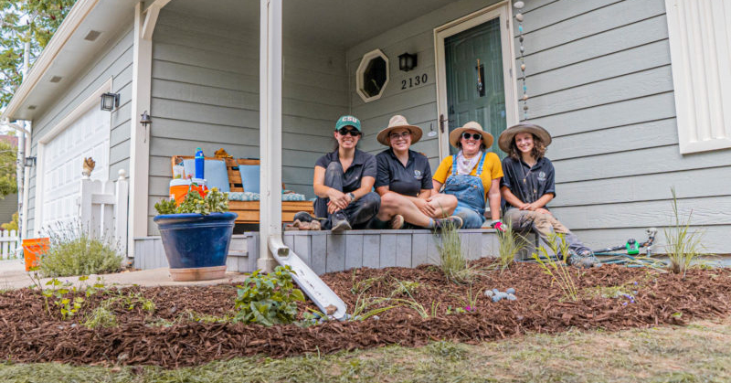Three people sit on a porch behind a neatly landscaped and mulched piece of yard beneath a gutter