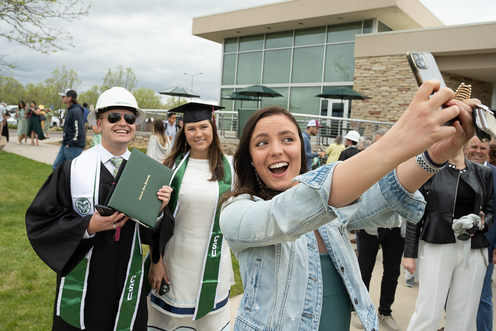 A young woman takes a selfie with two CSU graduates, one man wearing a hardhat and a woman wearing a graduation cap.