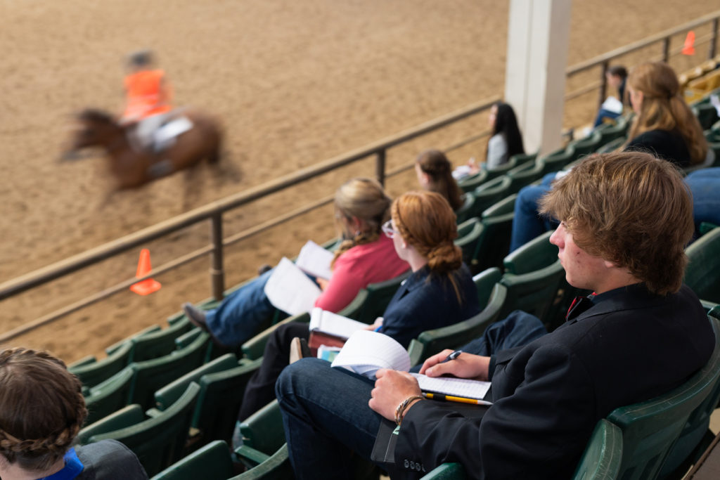 Young people sit in equine arena chairs taking notes, judging horsemanship
