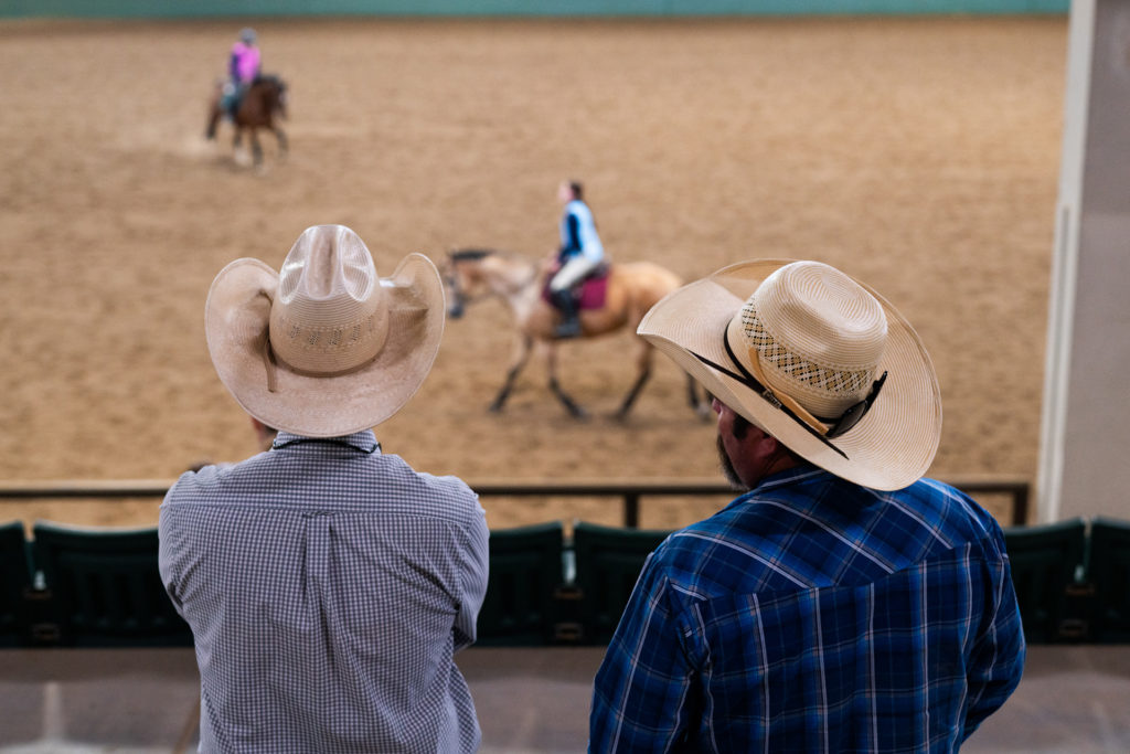Two men in western shirts and cowboy hats watch horse riders in arena