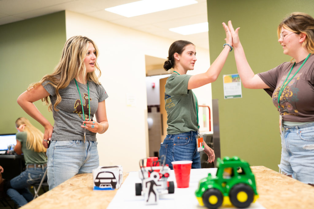 Two young women high five as a third speaks to them overlooking a table with a toy tractor, two red plastic cups and paper cutouts representing a car and people