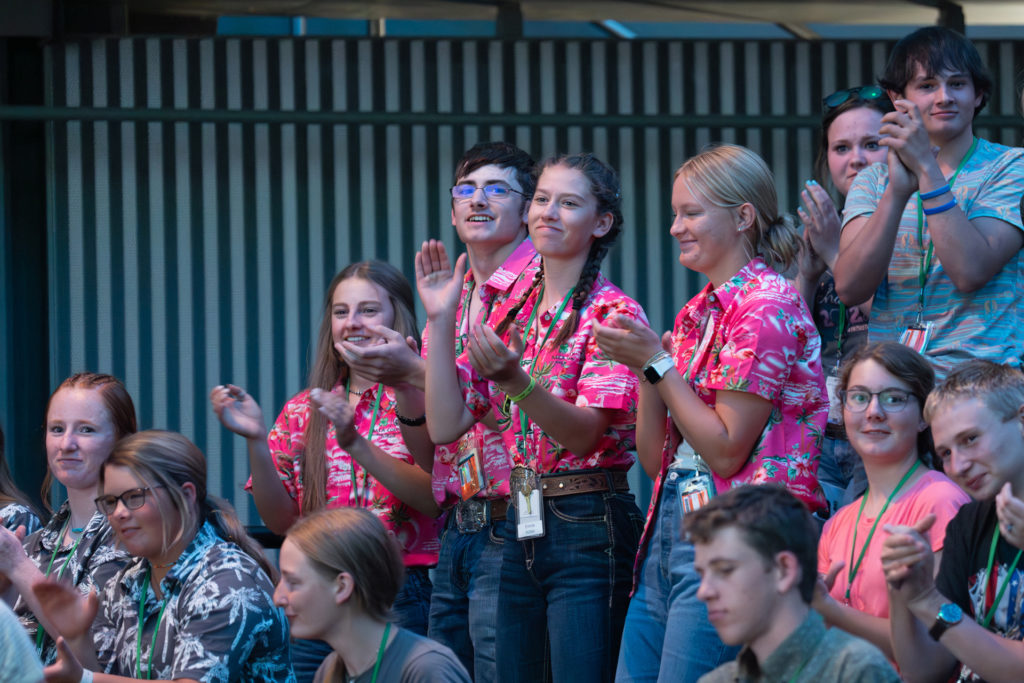 Young people dressed in matching, colorful shirts stand and applaud among seated crowd